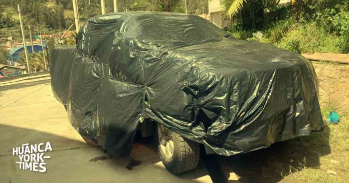 Car fully covered with a dark tarp parked outdoors beside a stone wall and greenery