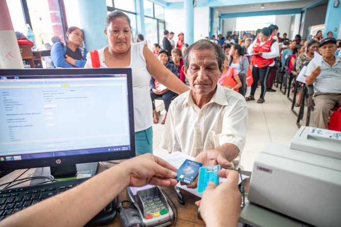 Older man seated at a desk as a clerk processes his payment with a card reader; people wait in line in a bright community hall far in the background.