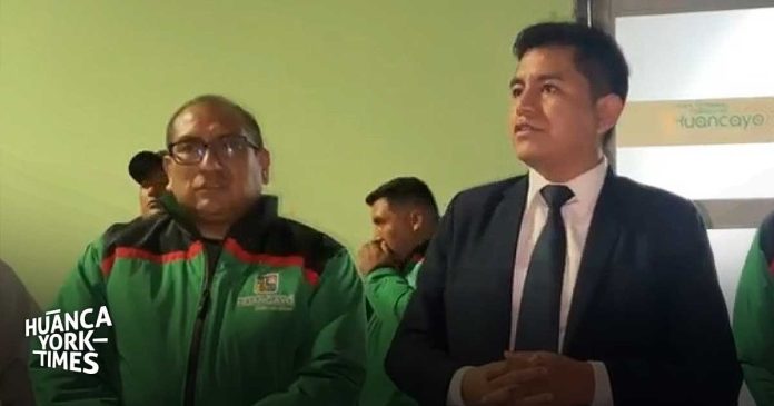 Two men at a press event: one in a green jacket with a logo, the other in a dark suit and tie; Huanca Times watermark.