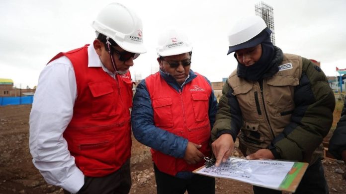 Three construction workers wearing hard hats and red vests examine a project blueprint on site.