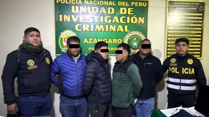 Group of five men standing in front of a green sign reading 'Unidad de Investigación Criminal' with two police officers nearby at a press event. Eye areas are blacked out for privacy.