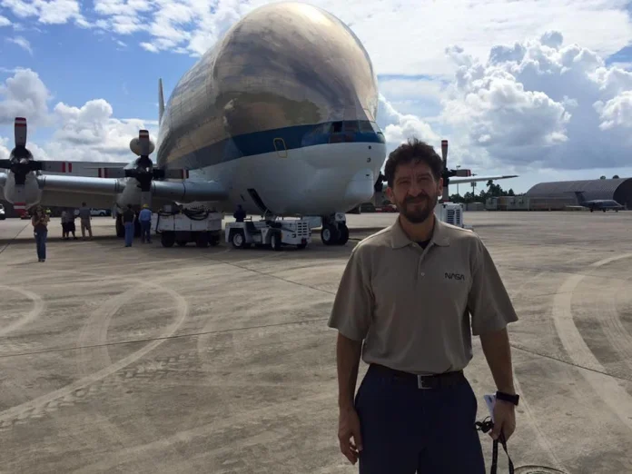 Man in a NASA polo stands on a tarmac beside a large silver propeller aircraft under a partly cloudy sky.