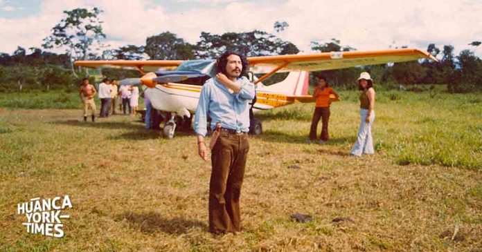 Man with long hair in a blue shirt stands in a field beside a small orange-and-white plane; people in background.