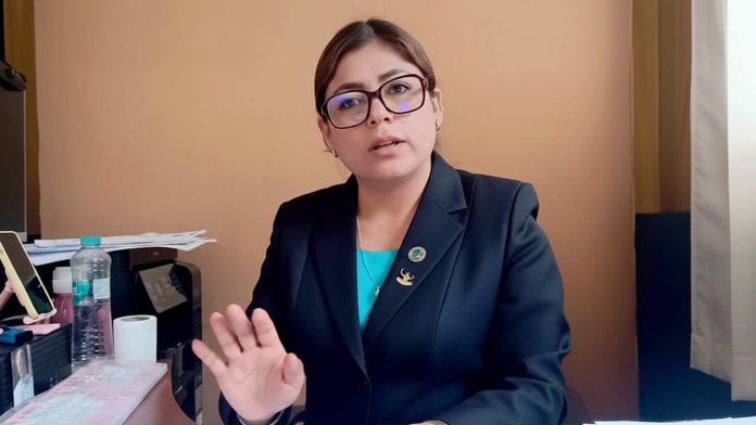 Professional woman in a dark blazer sitting at a desk, gesturing with an open hand toward the camera while speaking to the viewer.