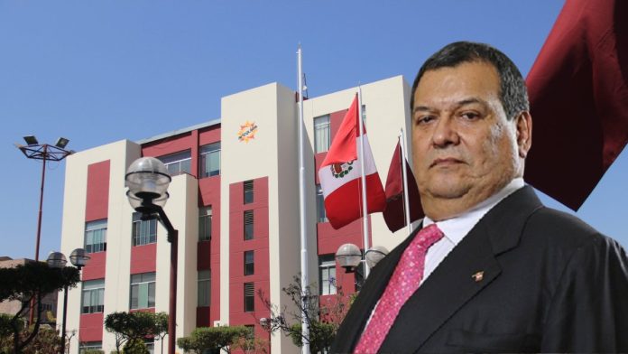 Man in a dark suit and pink tie stands before a modern red-and-cream government building with flagpoles.