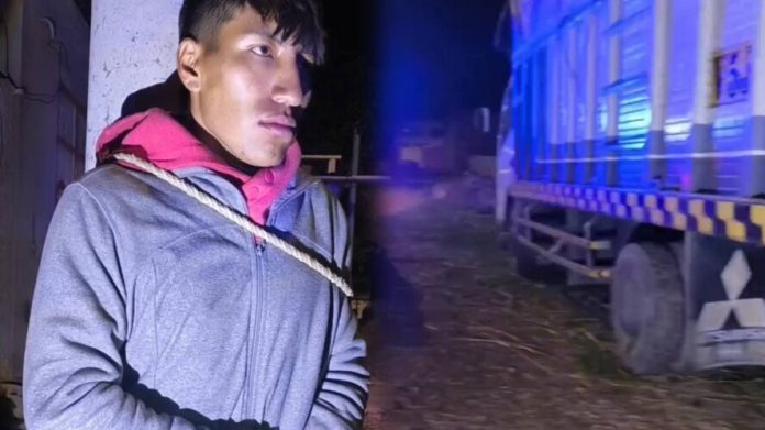 Man in a gray jacket and pink hooded scarf stands beside a rope tied across his chest, near a blue-lit train at night.