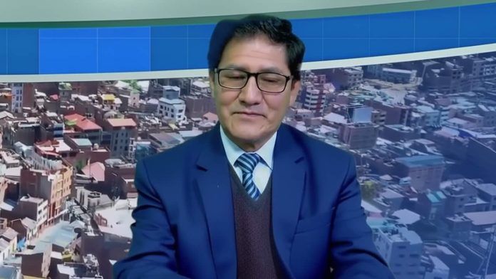 Male news presenter in a blue suit and striped tie sits at a desk with a cityscape backdrop behind him.