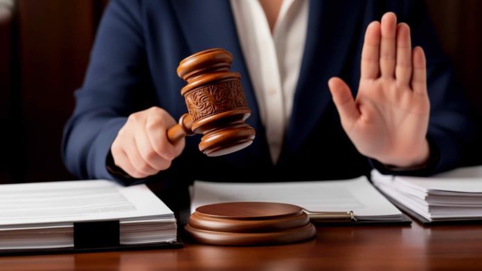 Judge's gavel raised for a strike on a desk, with a person in a suit holding it and a raised palm signaling pause or stop.