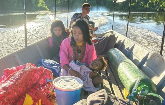 Woman in pink holds a child with oxygen tubing on a crowded boat drifting along a river.