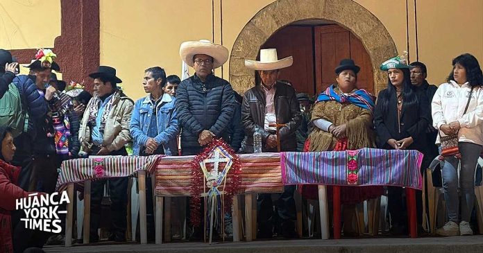 Group of people in traditional clothing and hats stand behind a long table draped with colorful woven fabrics at a cultural ceremony.