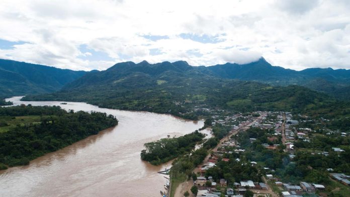 Aerial view of a muddy river winding through a green valley with a town along its banks and forested hills in the distance.