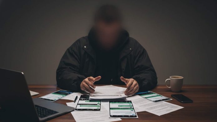 Hooded figure at a desk cluttered with papers and several smartphones displaying messages, plus a laptop and coffee mug nearby.