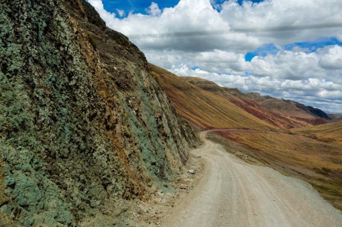 Winding dirt road beside a textured rocky cliff on the left, with rainbow-colored hills and a cloudy blue sky ahead.