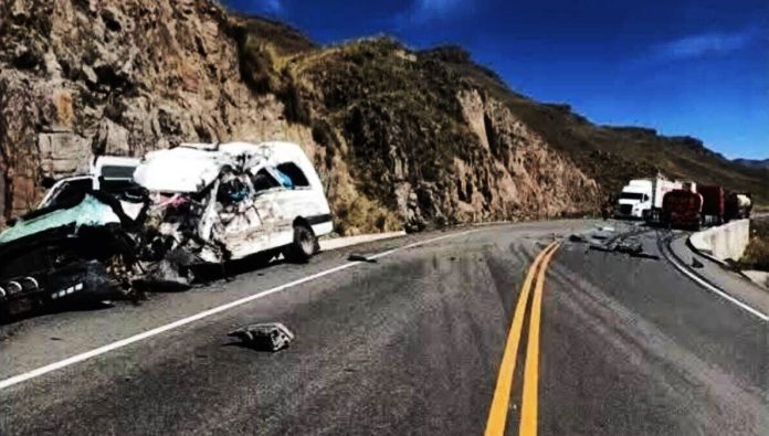 Two severely damaged vans sit on a winding mountain road with debris scattered, a rocky hillside on the left and a clear blue sky above.