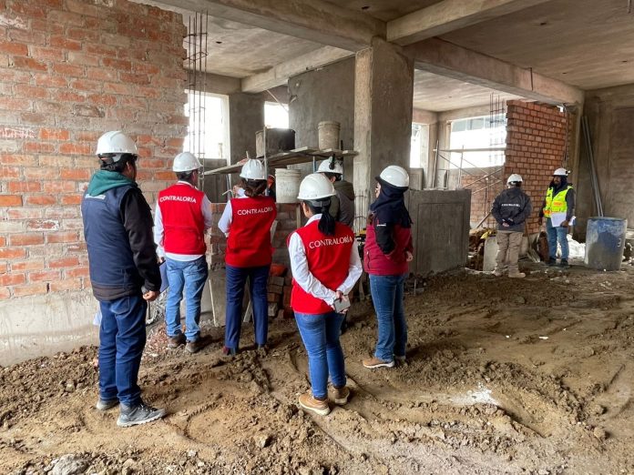 Group of inspectors in white helmets and red Contraloría vests listening to a briefing at an unfinished construction site in a concrete building.