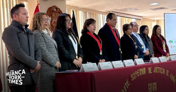 Nine officials stand behind a long table with name placards at a formal award or recognition ceremony; some wear medals and red scarves, with a Huanca York Times logo in the corner.