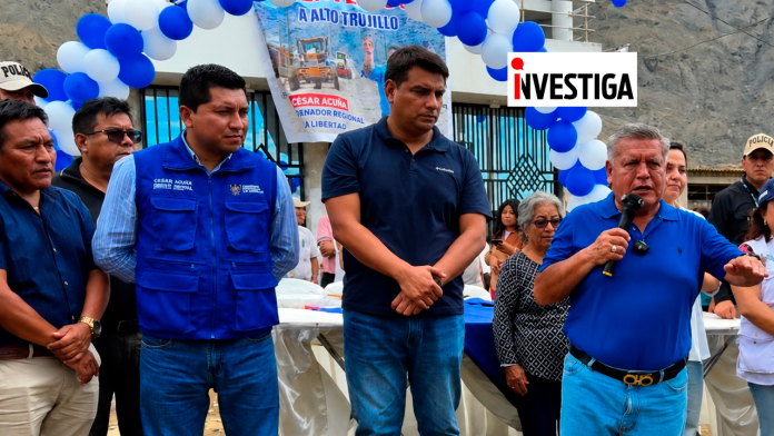 Public official speaks into a microphone at an outdoor event with blue balloons and a banner in the background, people listening nearby