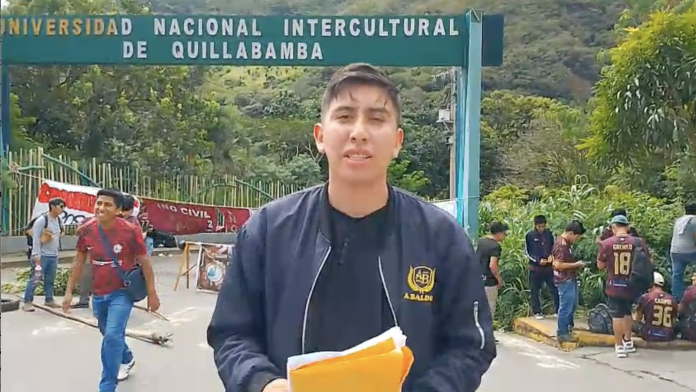 Student stands center frame, holding orange folders, at a university entrance with people nearby and greenery in the background.