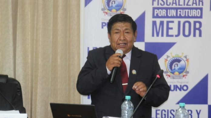 A man in a dark suit and red tie speaks into a handheld microphone at a conference table, with banners behind him.