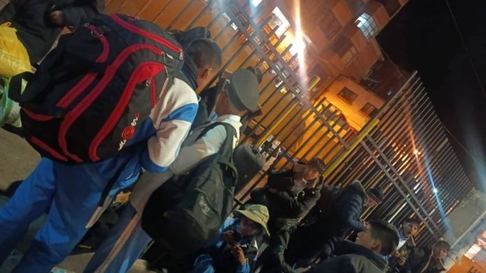Group of people with backpacks gathered along a tall metal gate at night, some sitting and others standing, waiting outside a fenced area.
