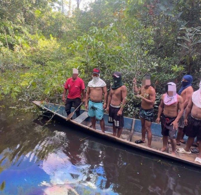Group of shirtless men with cloth masks standing along a narrow boat in a muddy jungle river.