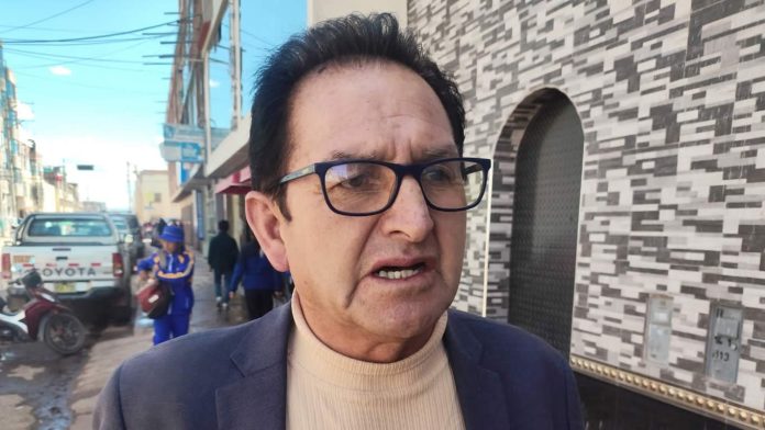 Man wearing glasses and a navy blazer on a busy street, with a tiled building facade in the background.