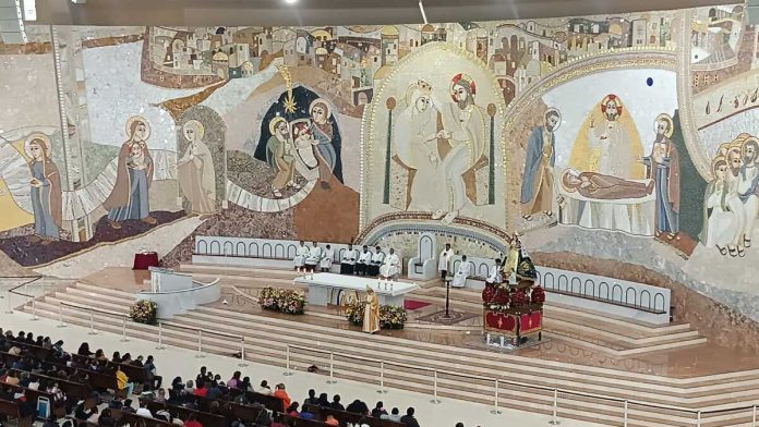 Inside a church with a large biblical-mosaic wall; clergy stand at the altar as worshippers fill the pews on stepped seating.