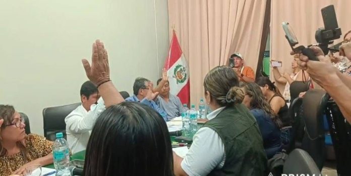 A group of people around a conference table, one person raising a hand, with a Peruvian flag in the background and reporters filming on the right side.