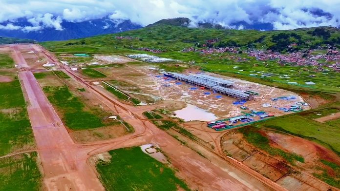 Aerial view of an airport construction site in a green valley, with a long white terminal under construction and a distant village on the hillsides.