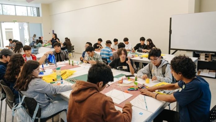 Group of students seated at long tables in a bright classroom, crafting with colored paper and markers.