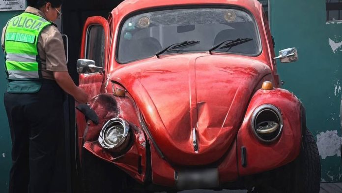 Police officer in a neon vest inspects a red, crumpled vintage car after a collision, focusing on the damaged front left side.