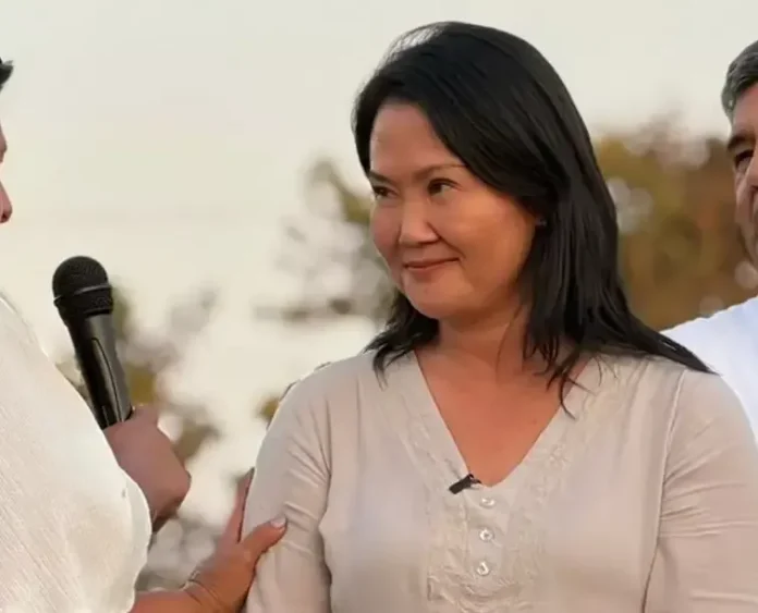 Smiling woman in a light beige blouse being interviewed outdoors; a microphone is visible near her.
