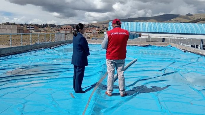 Two people inspect a blue rooftop pool cover; one person wears a red vest labeled 'Contraloría' and points toward the area.