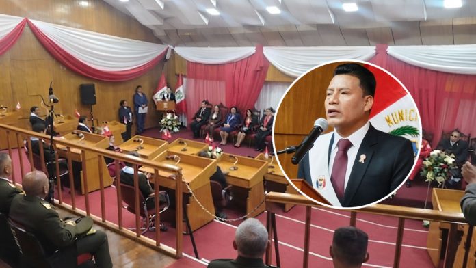 Formal council chamber ceremony with officials seated, red-and-white drapes, and flags in the background; inset shows a man in a suit speaking at a microphone.