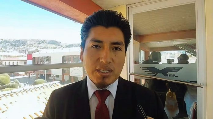 A man in a dark suit and red tie speaks to the camera on a balcony with glass doors behind him.