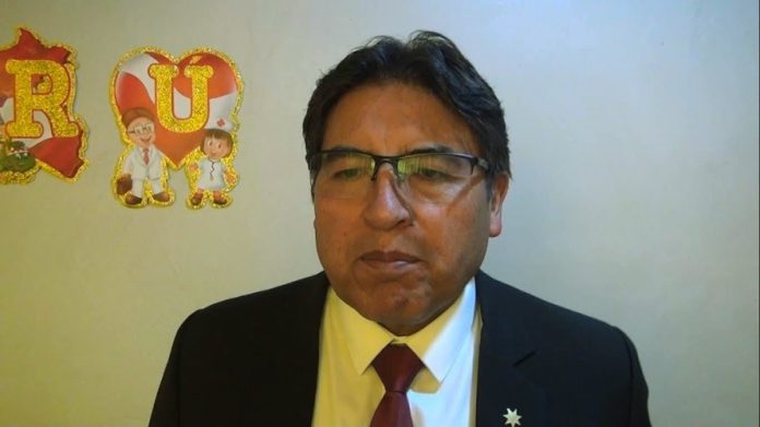 Man in a dark suit and red tie wearing glasses, standing indoors with decorative red-and-gold letters on the wall behind him (R U A).