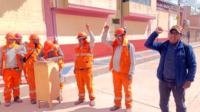 Group of construction workers in orange safety gear outdoors, one man signs a document at a wooden podium while others raise their fists in solidarity.