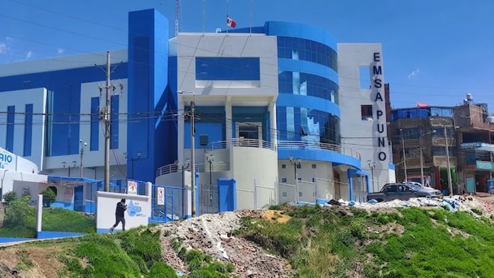 Modern blue-and-white hospital building with a tall vertical EMSAPUNO sign, gated entrance, and a person walking near rubble on a sunny day.