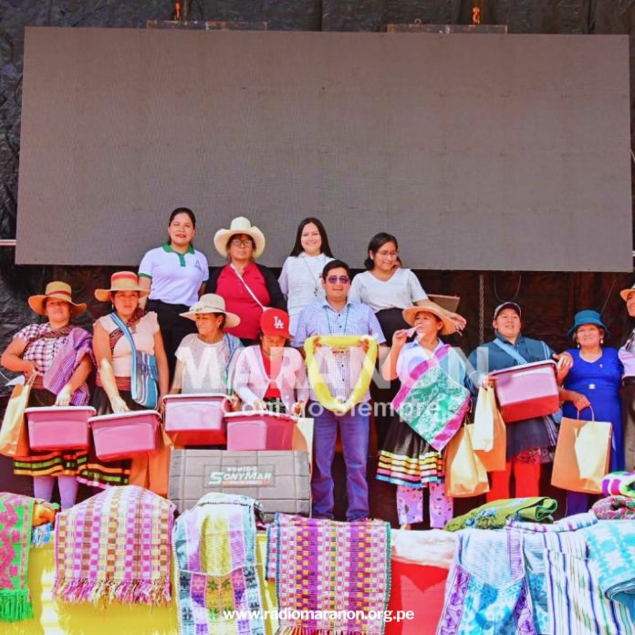 Group of men and women in traditional Andean clothing posing on a stage with colorful woven blankets and gift bags in front of them.